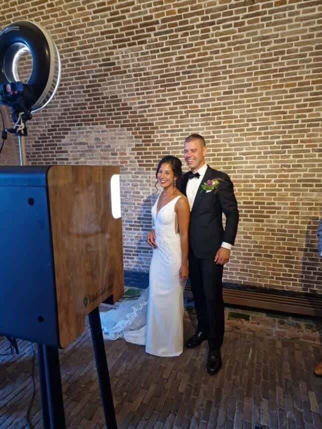 Gekleed bruidspaar maakt een stijlvolle foto bij een fotobooth tijdens hun bruiloft, met een bakstenen muur als achtergrond. De fotobooth legt romantische en persoonlijke momenten vast op het trouwfeest.