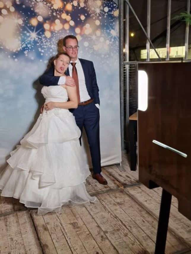 Bruidspaar poseert speels voor een photobooth op hun bruiloft met een sfeervolle achtergrond vol lichtjes. De fotobooth zorgt voor een leuke en spontane fotosessie tijdens het trouwfeest.