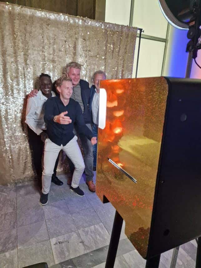 Groep vrienden staat lachend voor een glimmende gouden achtergrond bij een photobooth tijdens een trouwfeest. De fotobooth voegt plezier en feestelijke interactie toe aan de bruiloft. Met professionele dslr camera
